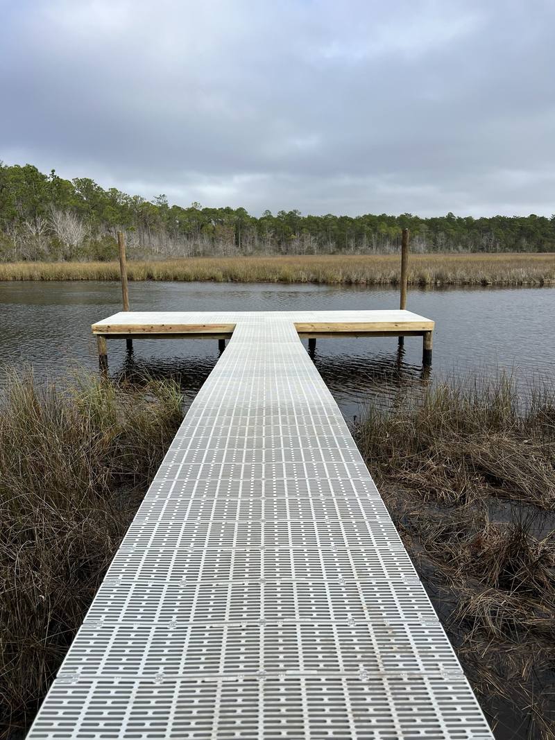 Long pier with light-colored grated decking crossing marsh grass to a T-shaped platform