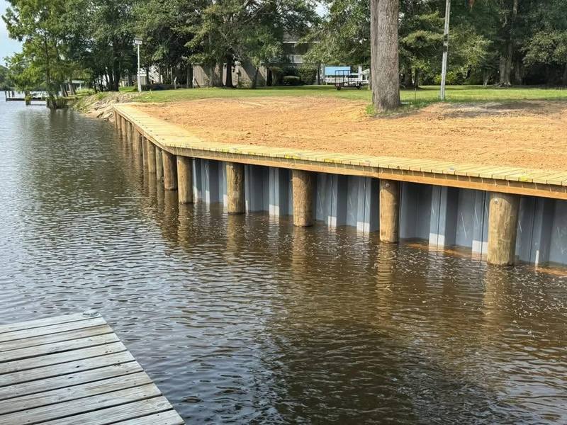 Long run of new vinyl bulkhead capped in wood along a pine-shaded bank, with a deck in the corner